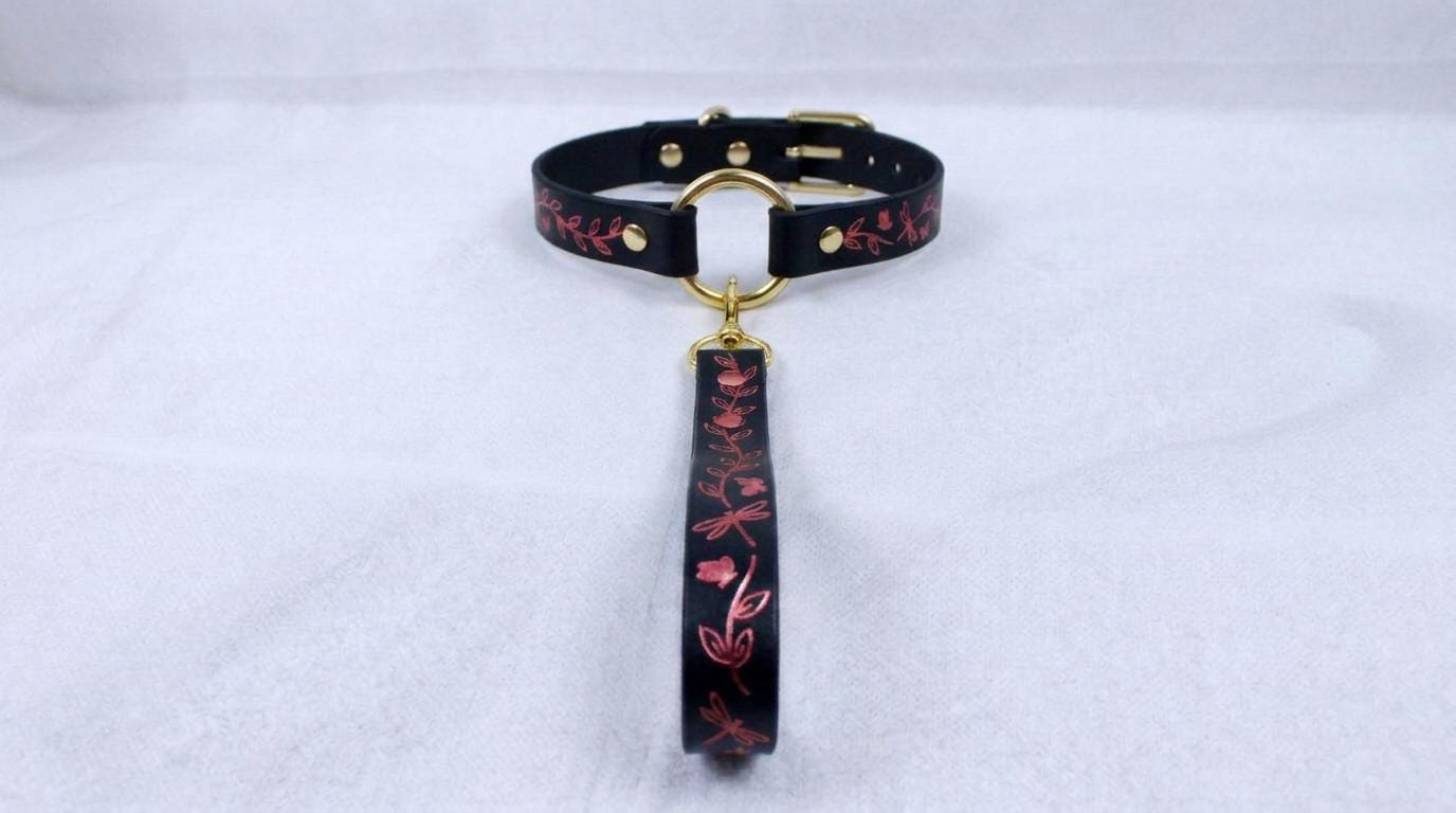 A black collar with a gold-colored buckle and a floral pattern on the strap is displayed on a white surface.