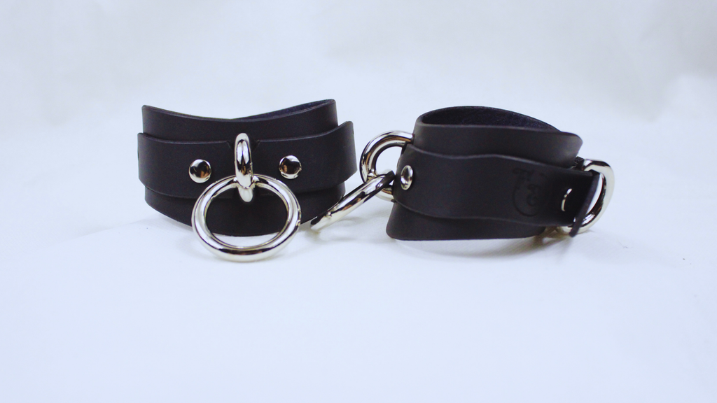 A pair of black leather choker straps with metal rings and buckles, displayed against a white background.