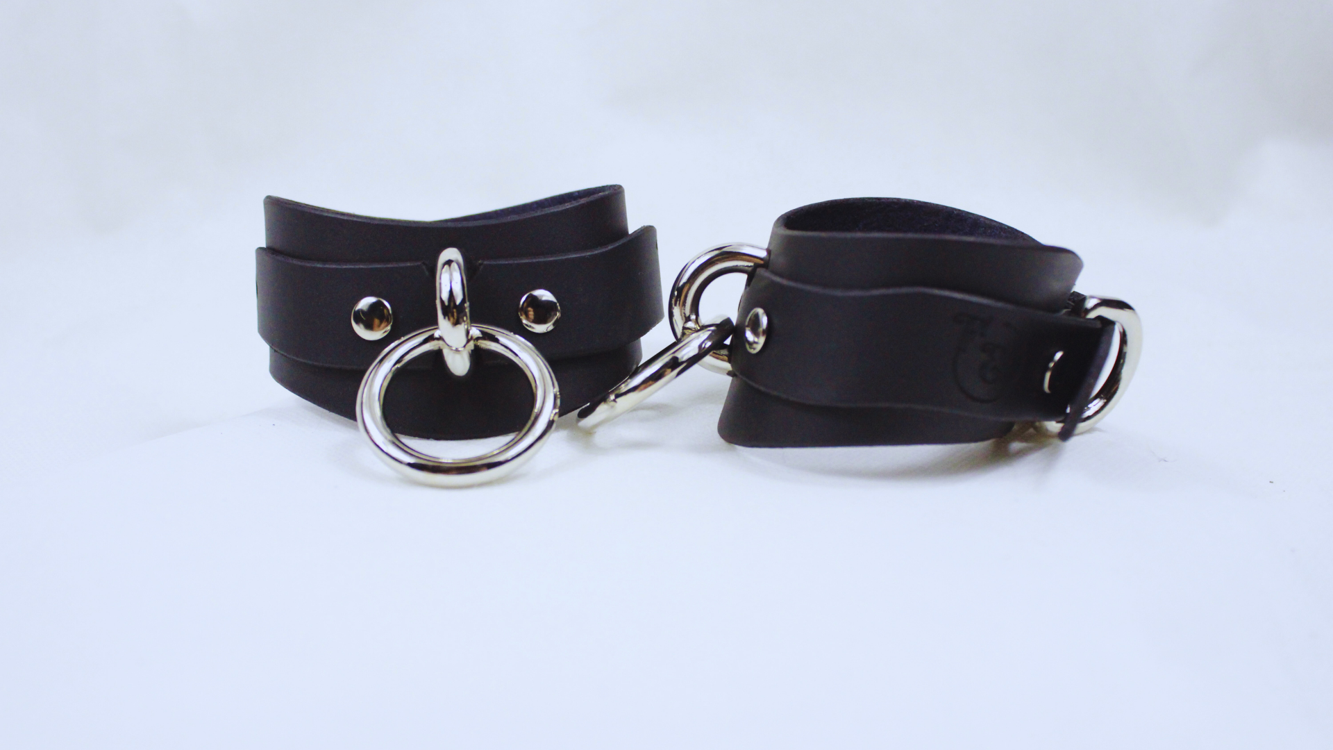 A pair of black leather choker straps with metal rings and buckles, displayed against a white background.