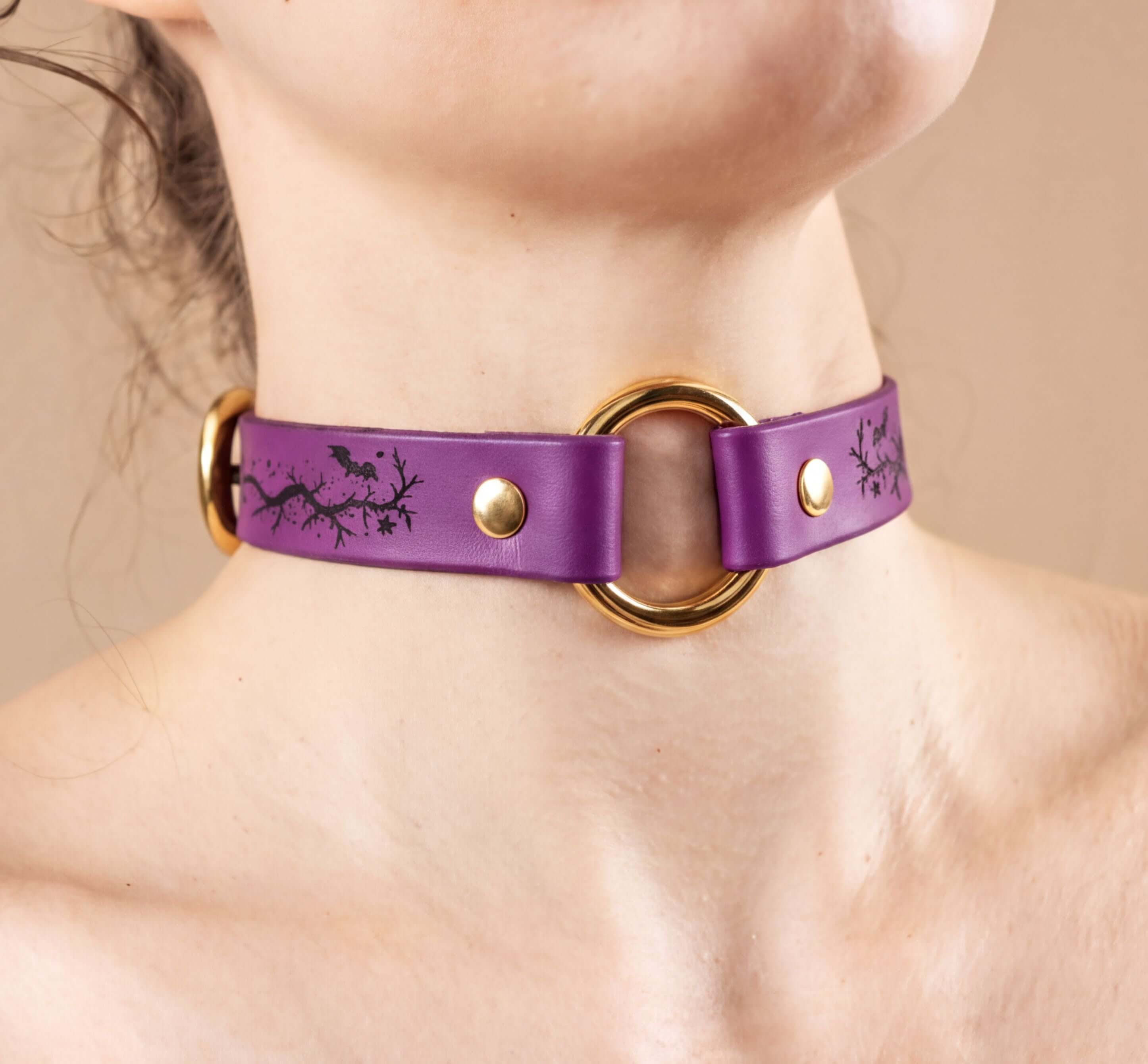 A close-up of a person's neck wearing a purple leather choker necklace with a gold ring and two gold buckles.
