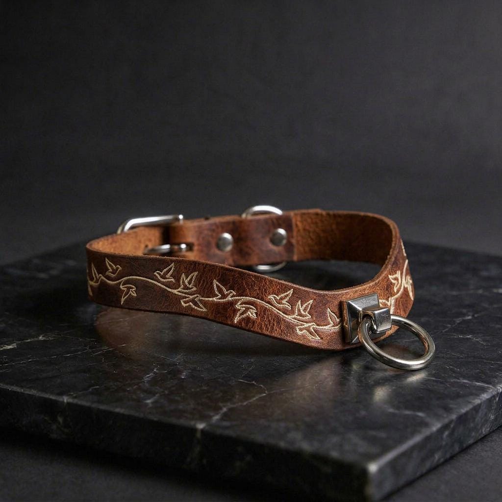 A brown leather collar with a metal ring and a leaf design on it, resting on a black marble surface.