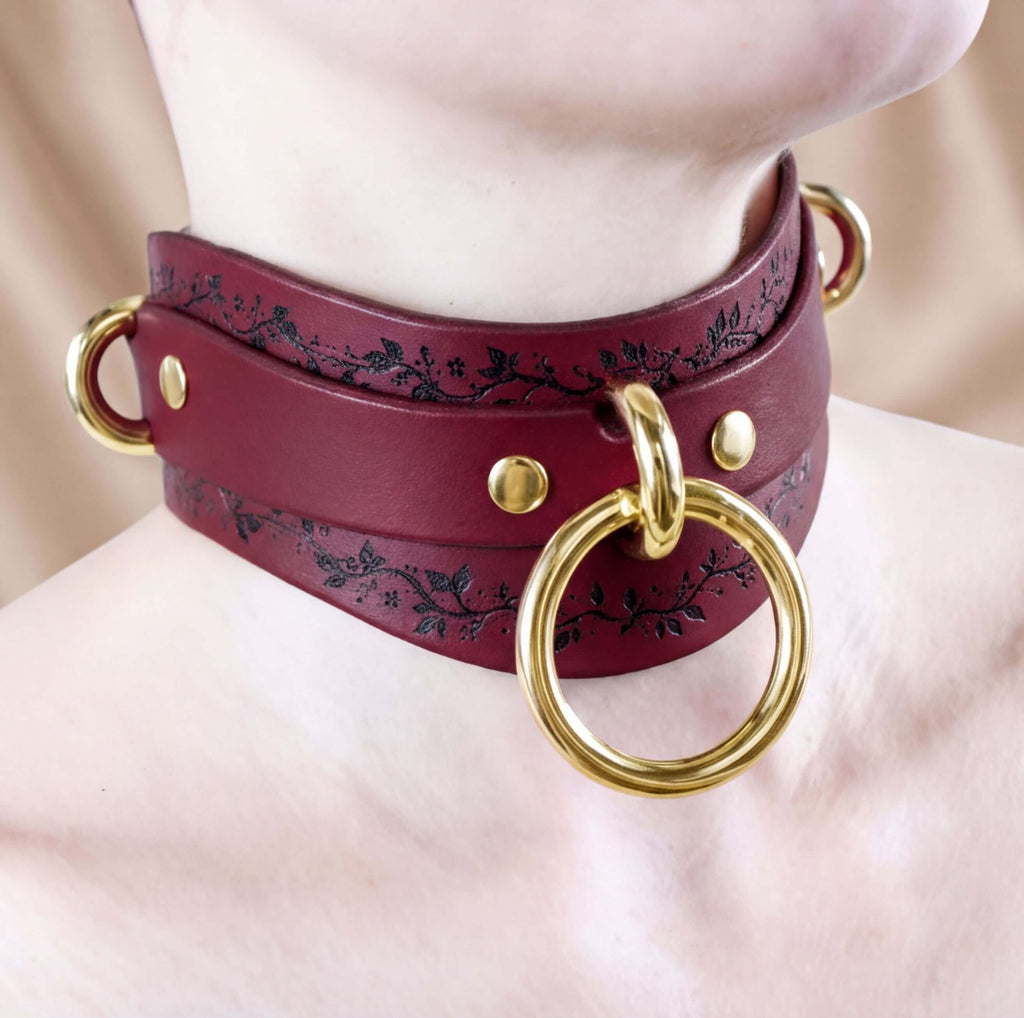 A close-up of a burgundy leather choker necklace with a gold ring and two gold rings on either side.