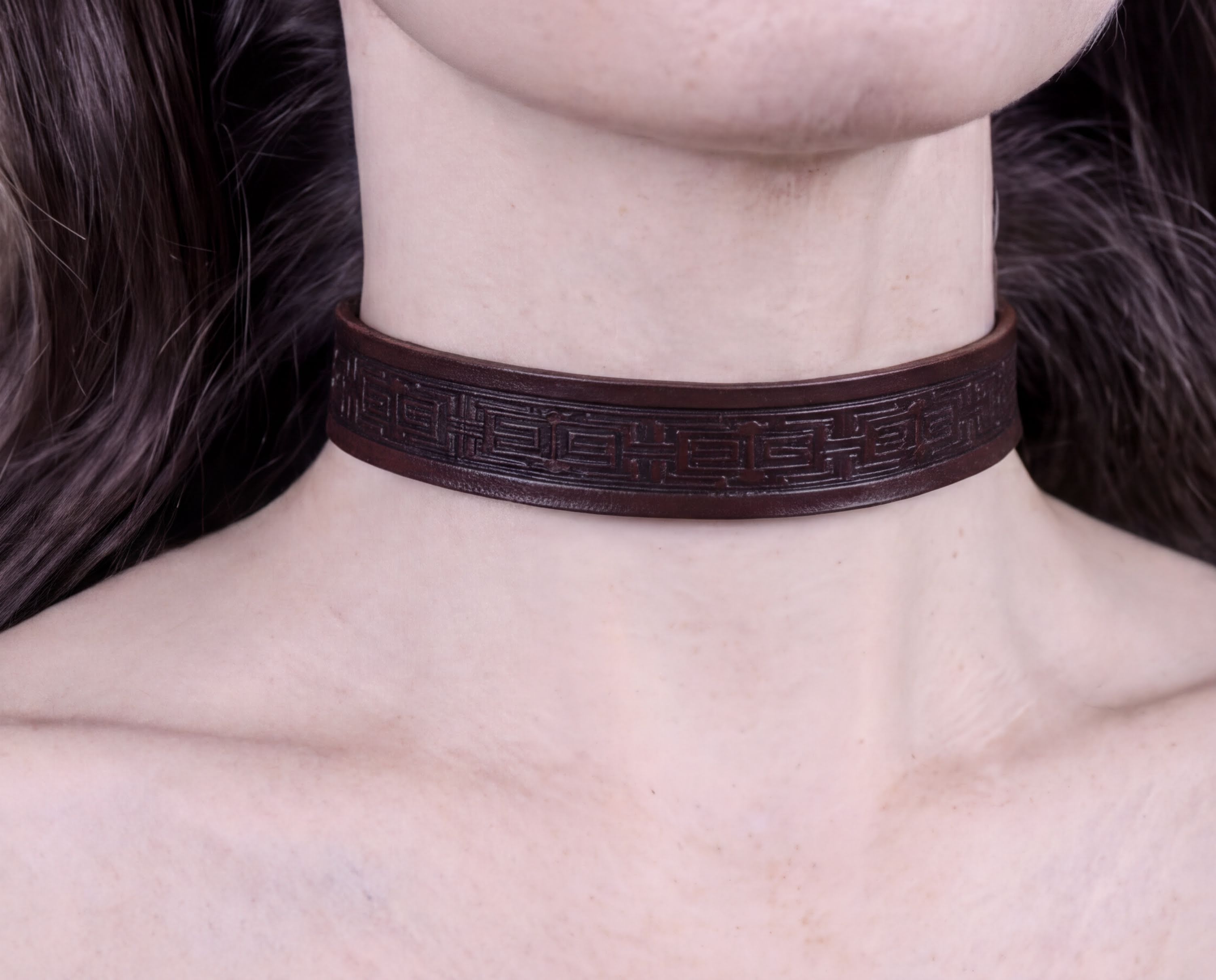 A close-up view of a person's neck and chest area wearing a dark brown leather choker necklace with an intricate geometric pattern.