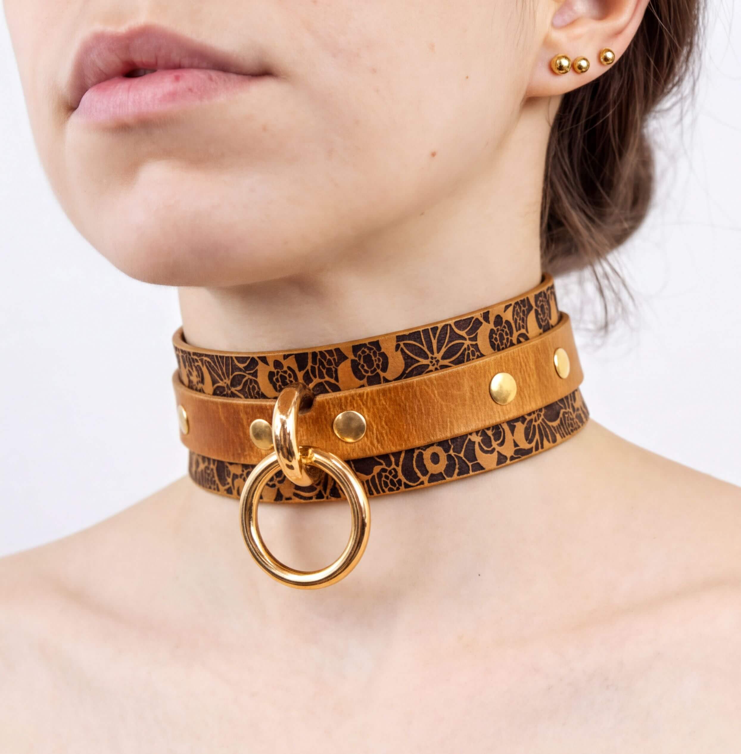 A close-up of a woman's neck and chest area wearing a choker-style leather collar with a gold ring and studs.