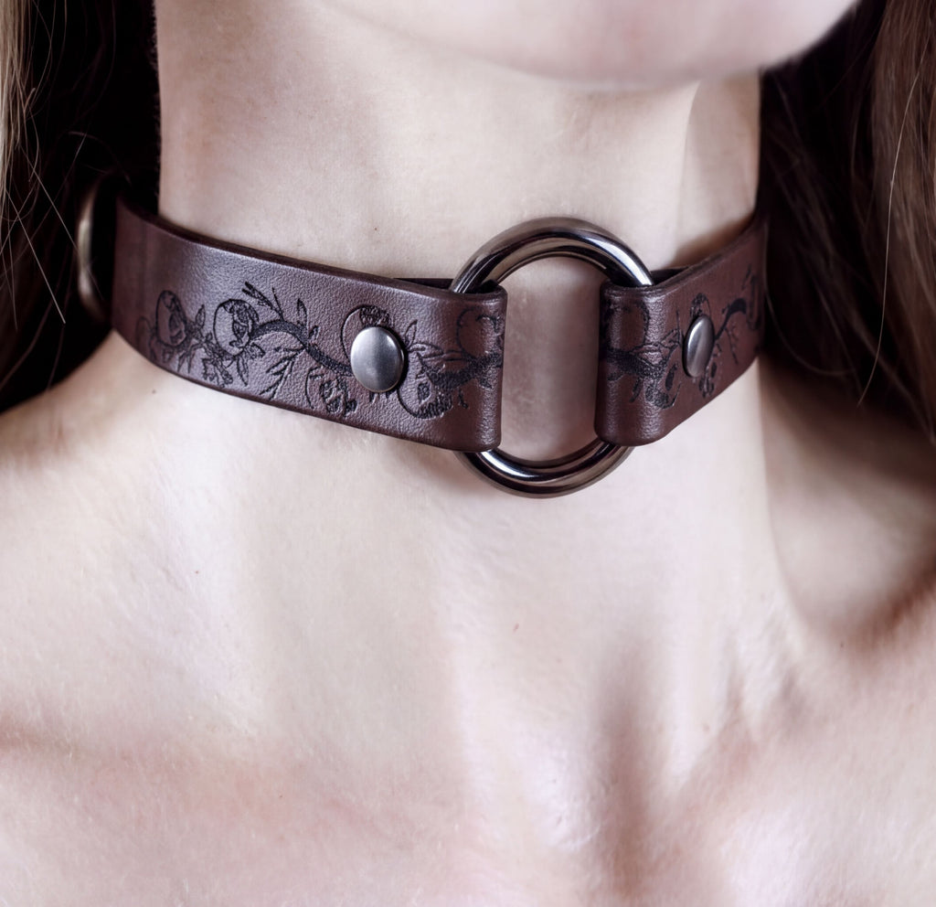 A close-up image of a woman wearing a brown leather choker necklace with a metal ring and two metal buttons.