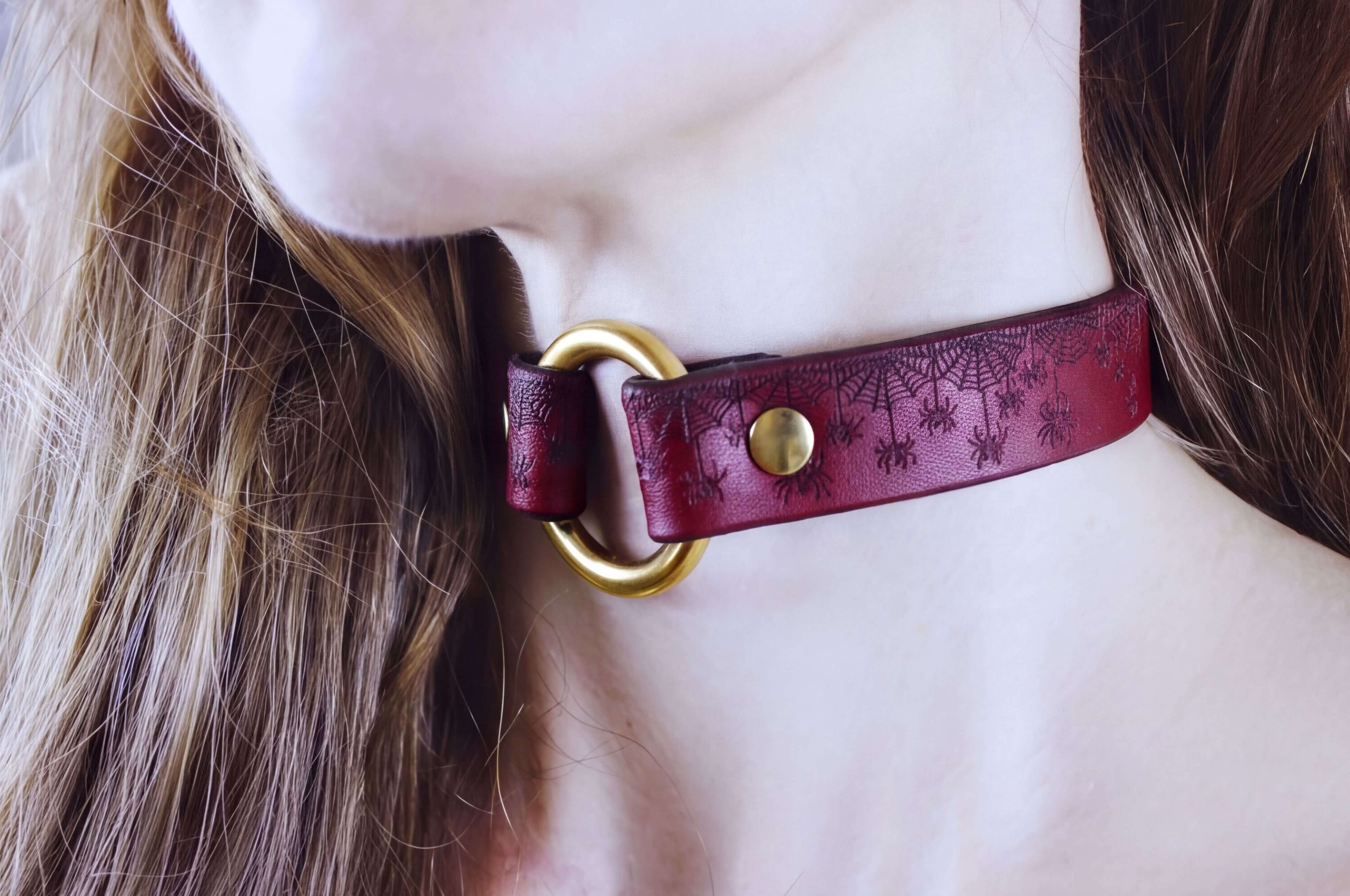 A close-up of a person's neck wearing a burgundy choker necklace with a gold ring and a spider web design.