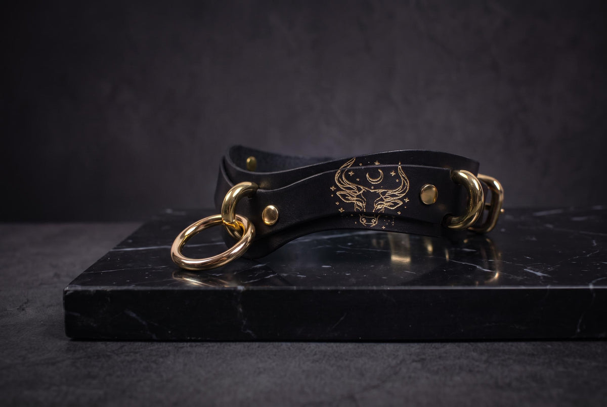 A black leather choker necklace with gold-colored metal rings and a design on the clasp, resting on a black marble surface.