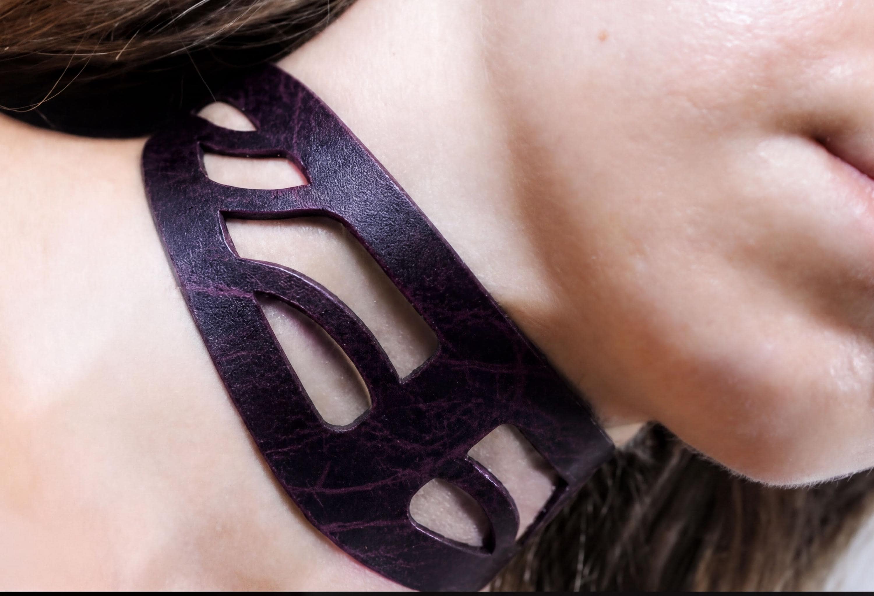 A close-up view of a person's neck and chin area, featuring a dark purple, leather-like necklace or choker with a unique, abstract cutout design.
