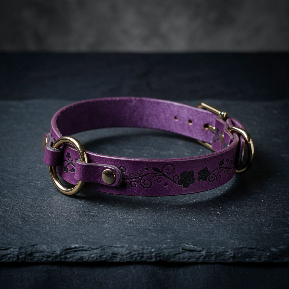 A purple leather collar with a floral design and metal rings is displayed on a dark surface.