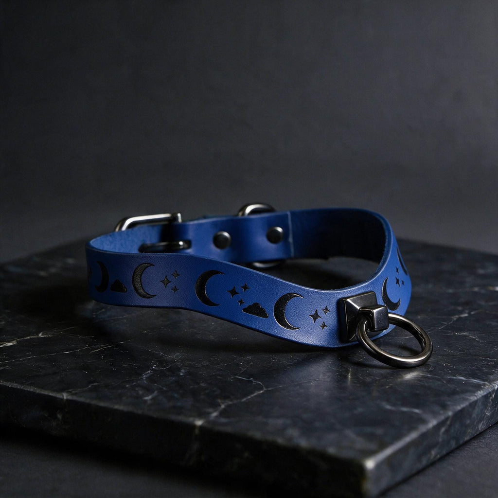 A blue leather collar with moon and star designs, featuring a metal ring and buckle, is displayed on a dark surface.