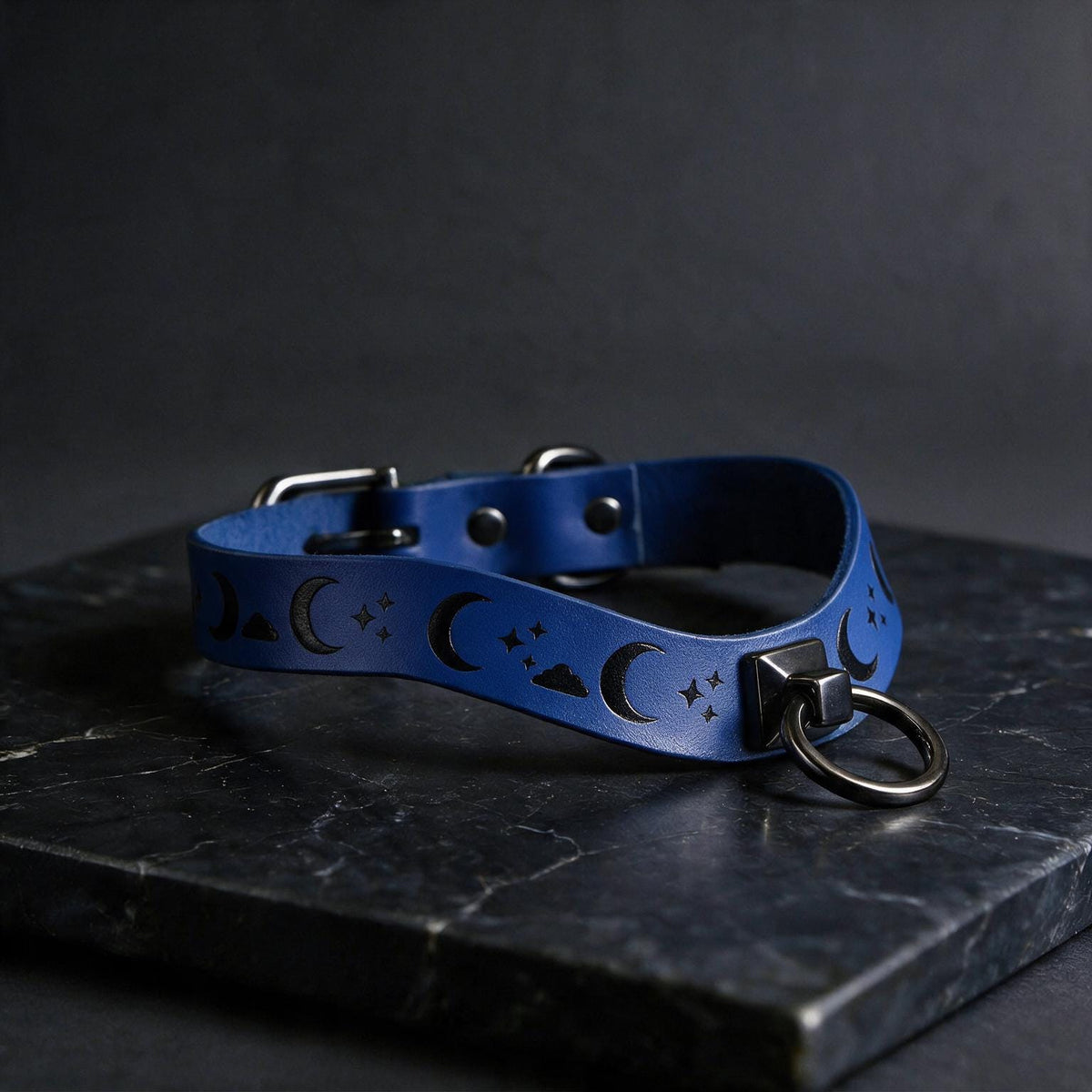 A blue leather collar with moon and star designs, featuring a metal ring and buckle, is displayed on a dark surface.