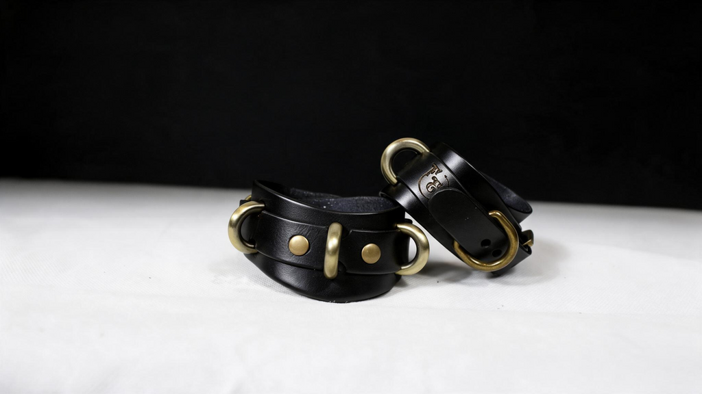 A pair of black leather cuffs with gold hardware, including buckles and clasps, are displayed against a black background.