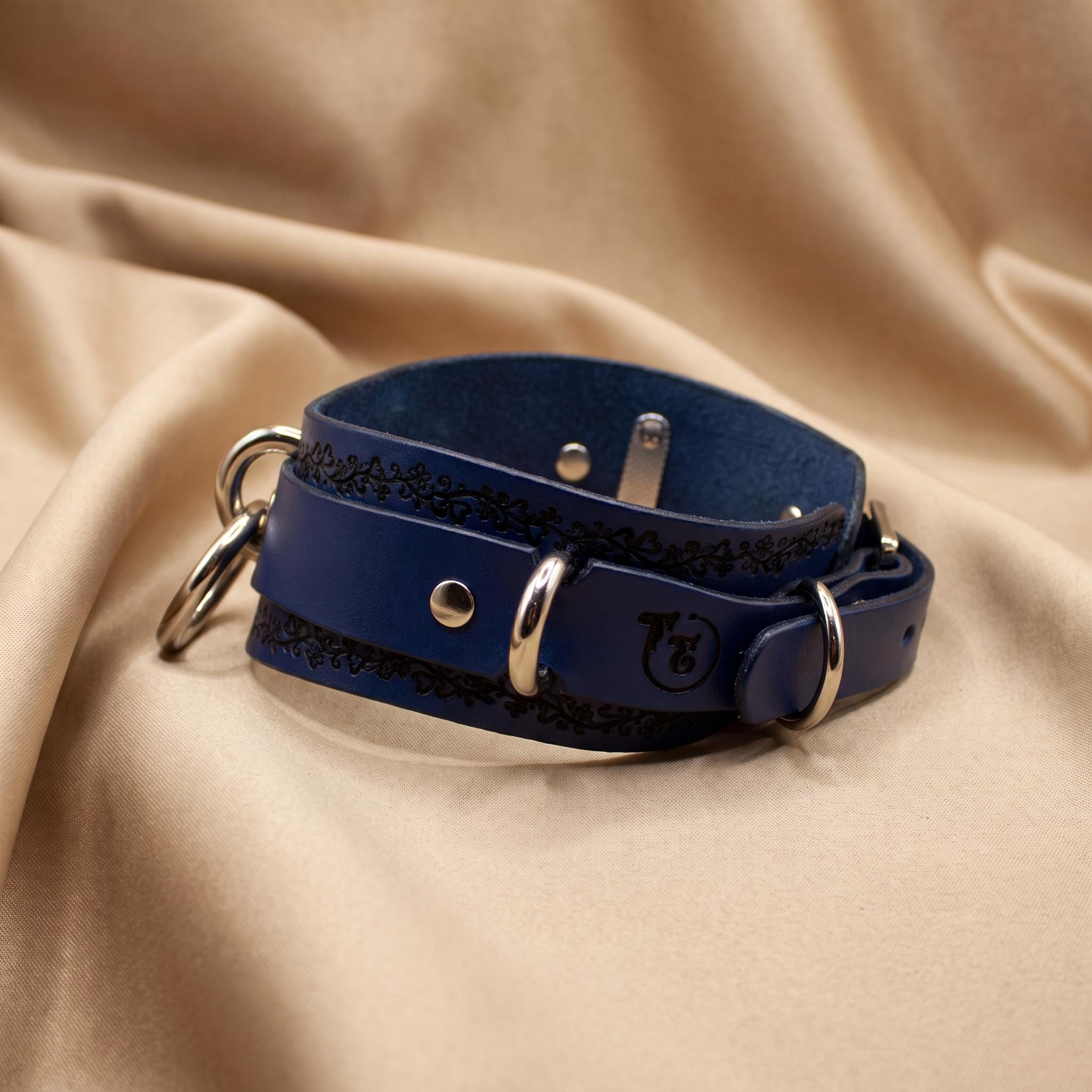 A blue leather collar with metal rings and a decorative pattern on the edges, resting on a beige fabric background.