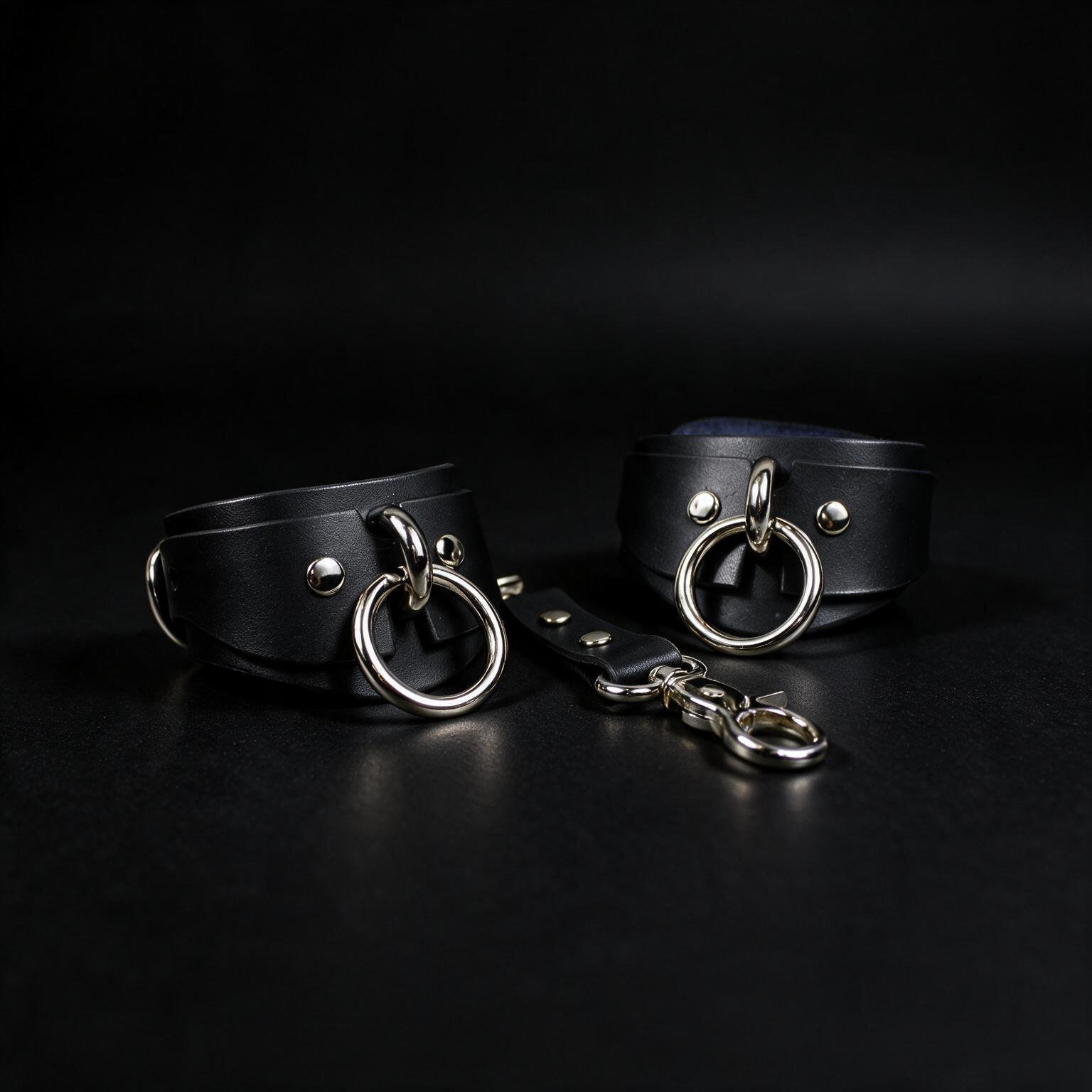 a pair of black leather choker straps with metal rings and buckles, placed on a dark background.