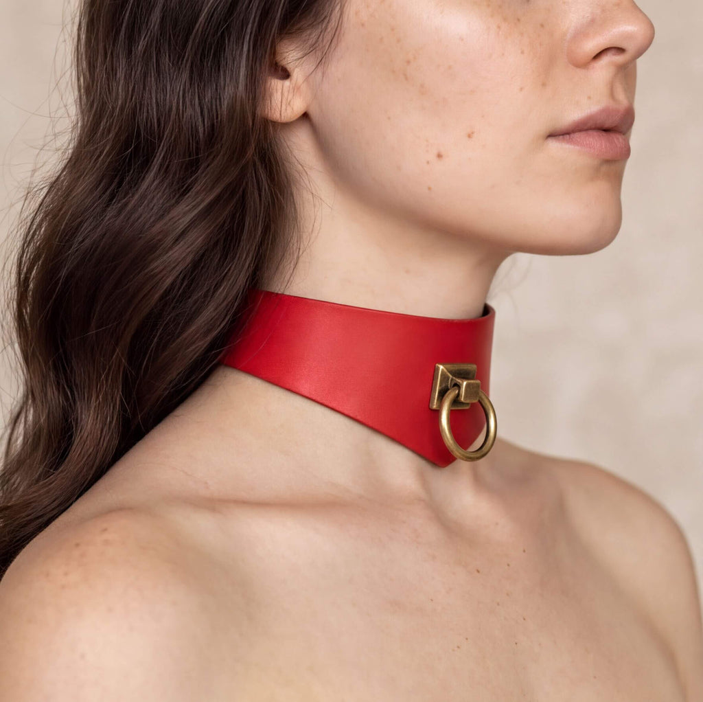 A close-up of a woman's neck and chest area wearing a red leather choker necklace with a gold-colored metal ring.