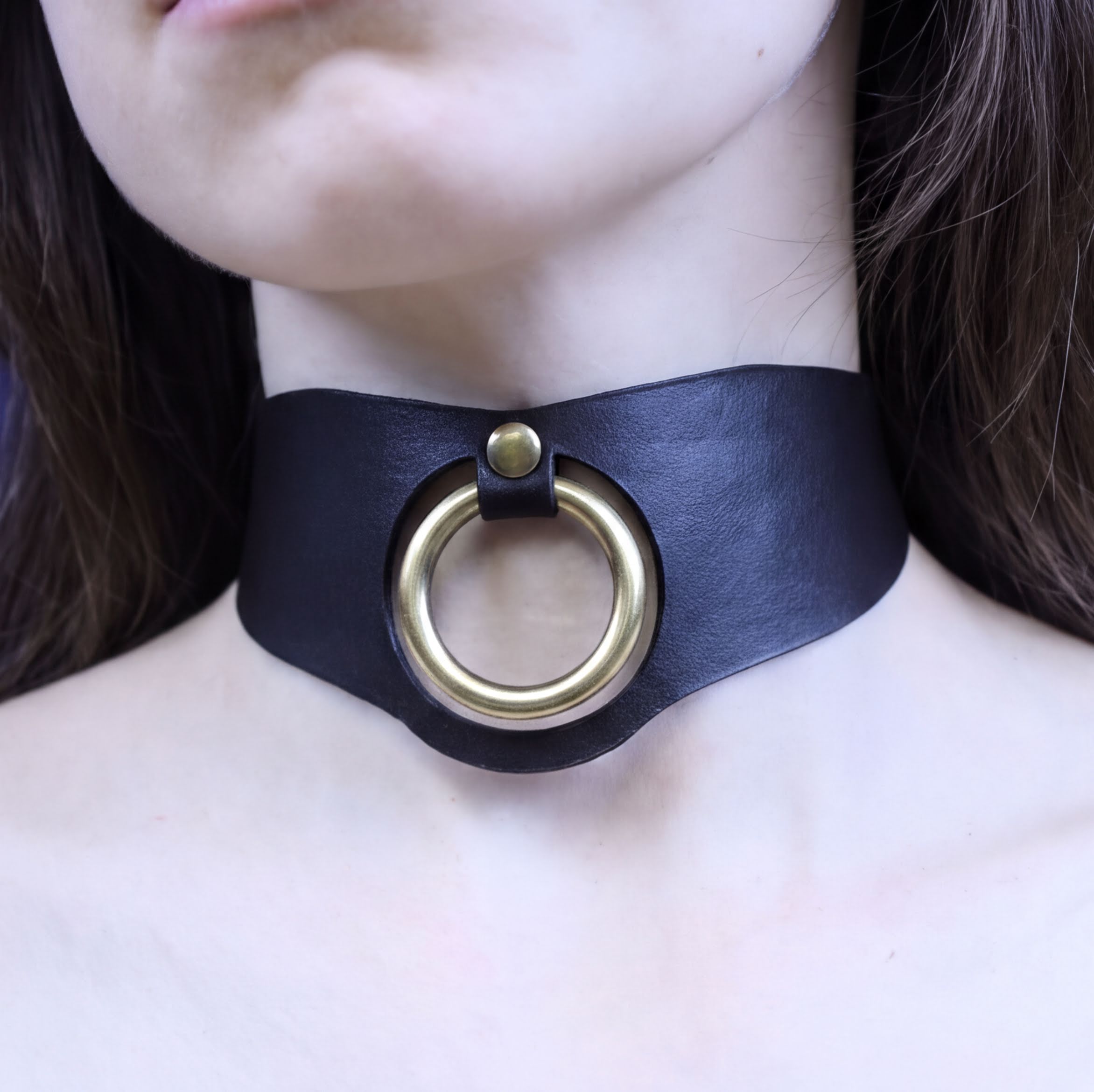 A close-up of a person's neck wearing a black leather choker necklace with a gold-colored circular ring.