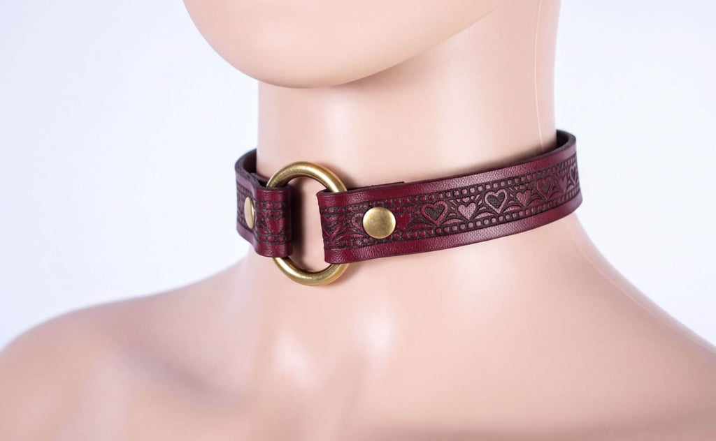 A close-up of a mannequin wearing a burgundy leather choker necklace with a gold ring and heart-shaped embellishments.