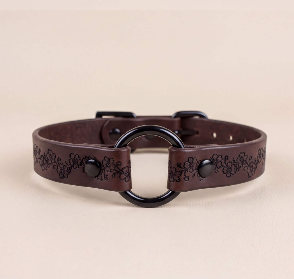 A brown leather collar with a black circular buckle and floral designs on the strap.