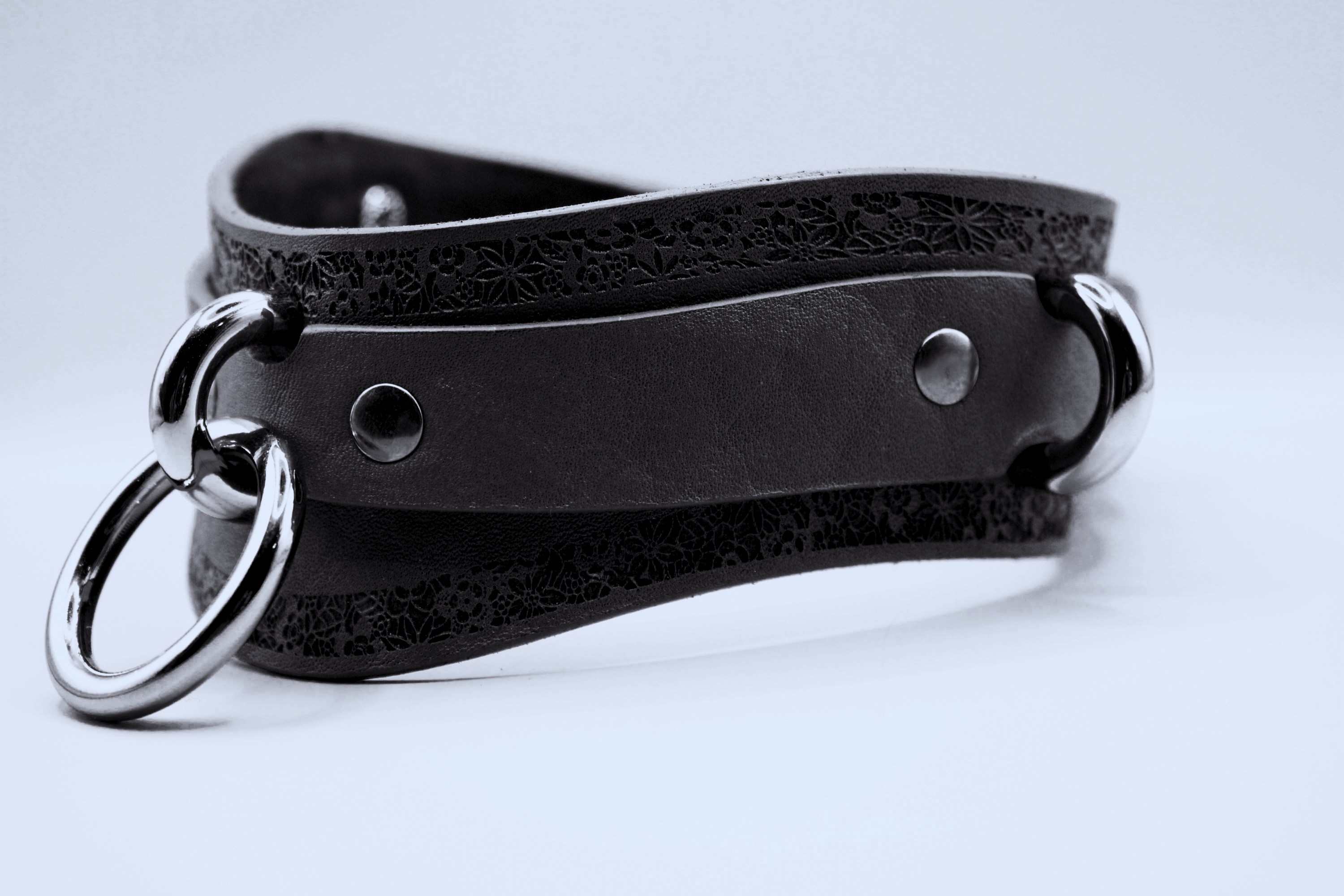 A close-up black and white image of a leather choker necklace with metal rings and a decorative pattern.