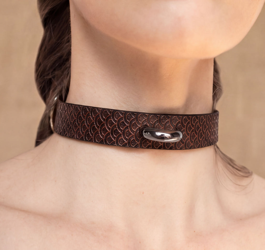 A close-up of a person's neck and chest area wearing a brown leather choker necklace with a metal clasp.