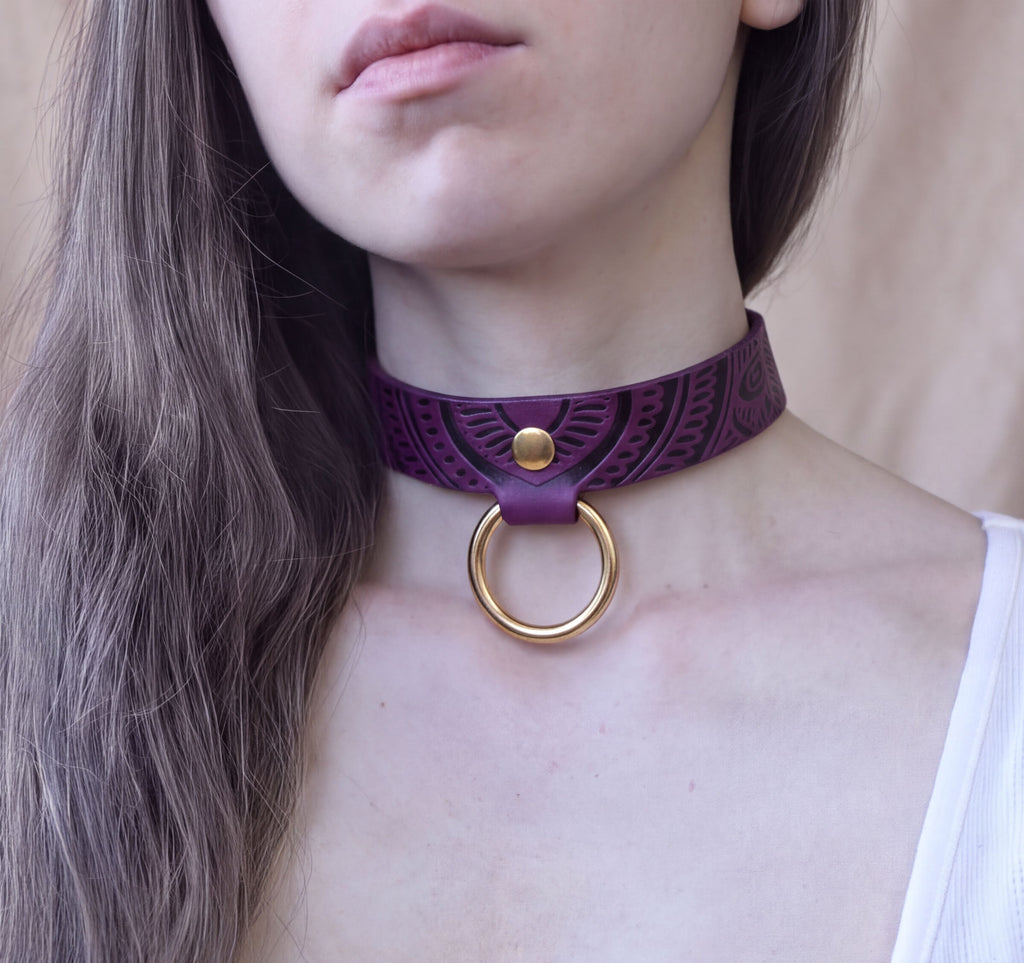 A close-up of a woman's neck and chest area, featuring a purple choker necklace with a gold ring.