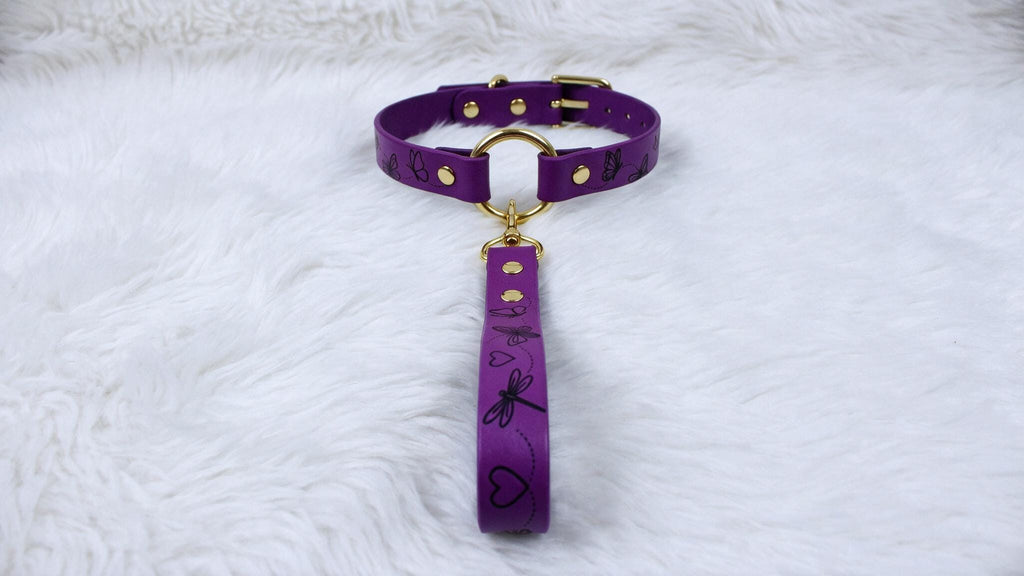 A purple dog collar with a gold buckle and handle is displayed on a white furry surface.