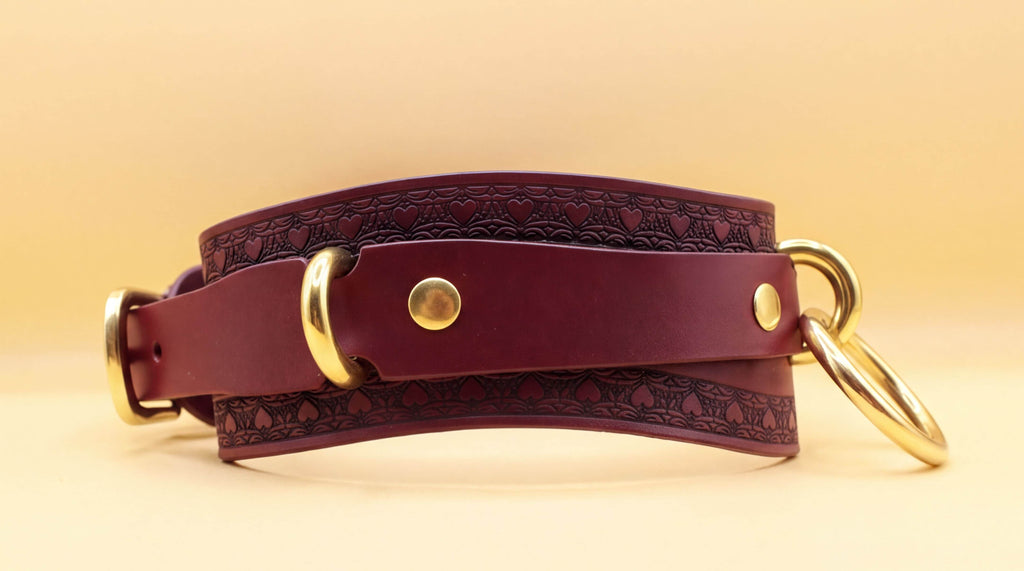 A burgundy leather choker with gold hardware, including two round buckles and two metal rings, is shown against a yellow background.