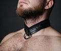 A close-up of a man's neck and chest area, with a black leather choker collar featuring metal rings and studs.