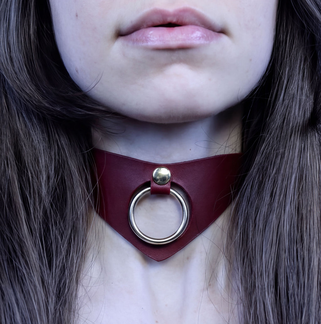 A close-up of a person's neck and chest area, featuring a burgundy-colored choker necklace with a circular metal ring.