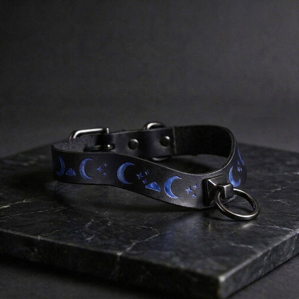A black leather collar with blue moon and star designs on it, resting on a dark surface.