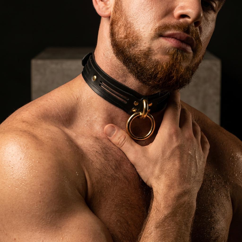 A close-up of a shirtless man with a beard wearing a black leather collar with a gold ring.