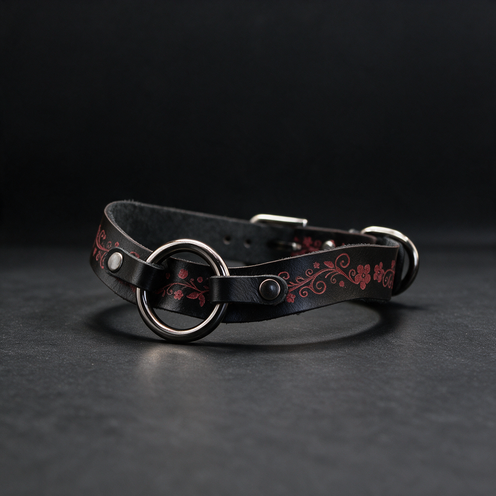 A black leather collar with a floral design and metal ring on the front, resting on a dark surface.