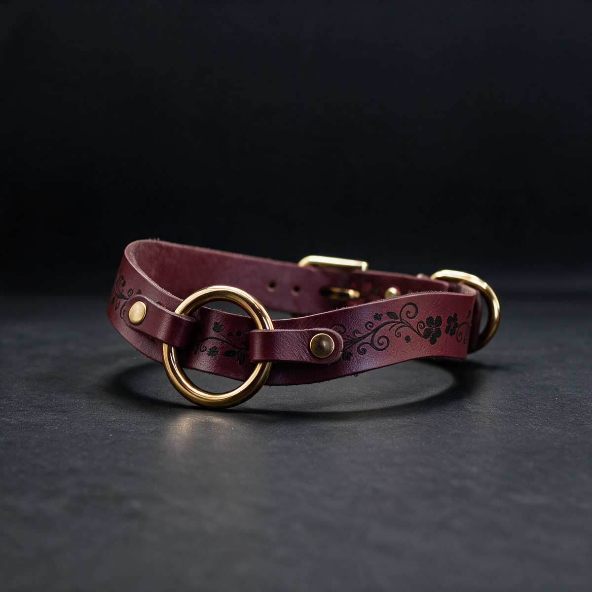 A burgundy leather collar with a gold-colored buckle and a floral design on the strap is displayed against a dark background.