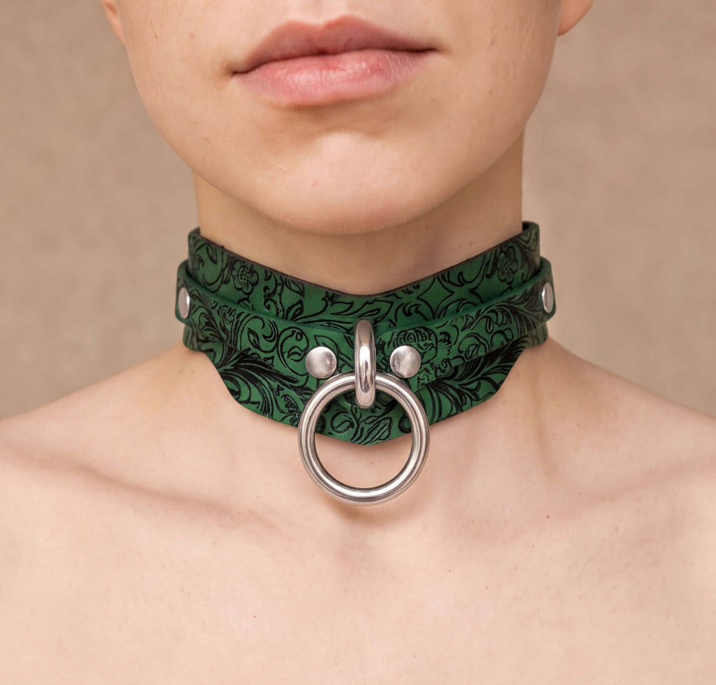 A close-up of a person's neck and chest area wearing a green choker necklace with a silver ring and metal studs.