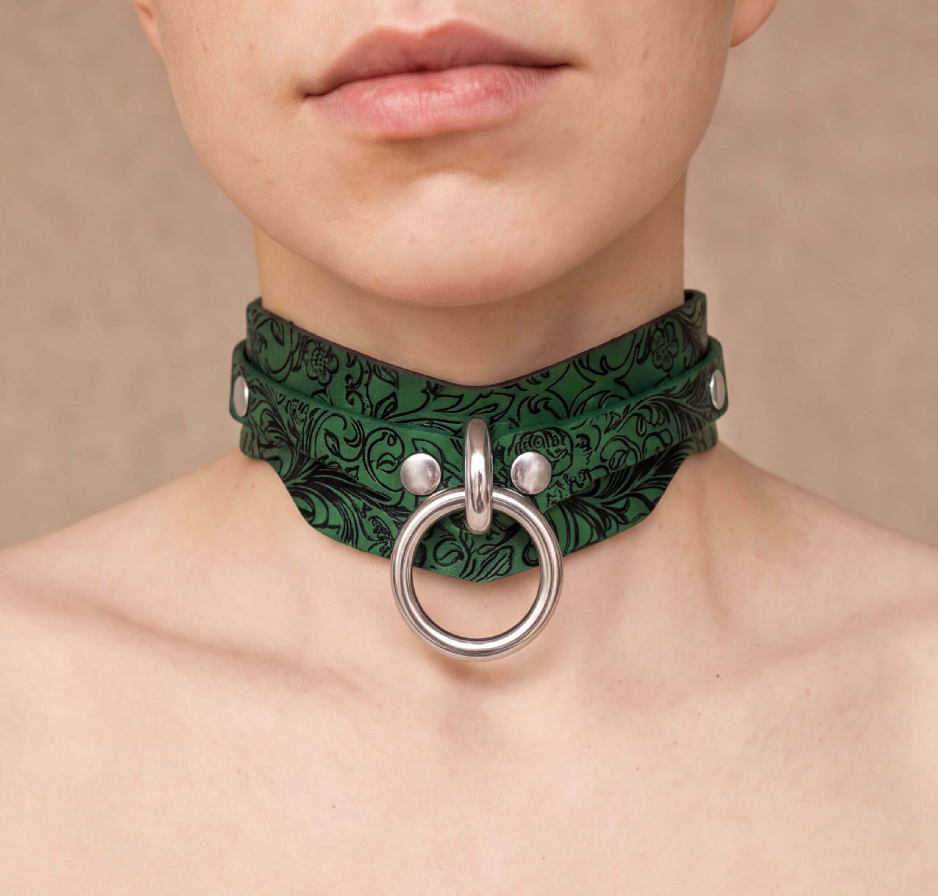 A close-up of a person's neck and chest area wearing a green choker necklace with a silver ring and metal studs.