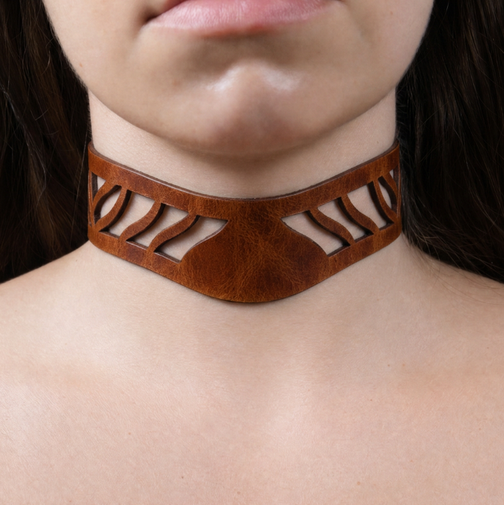 A close-up of a person's neck and chest area wearing a brown leather choker necklace with a unique cutout design.