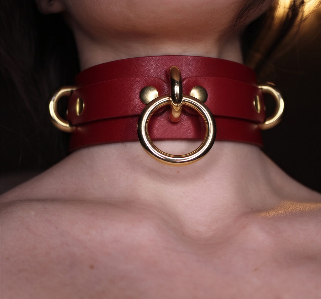 A close-up of a person's neck wearing a red leather choker necklace with gold-colored metal rings.