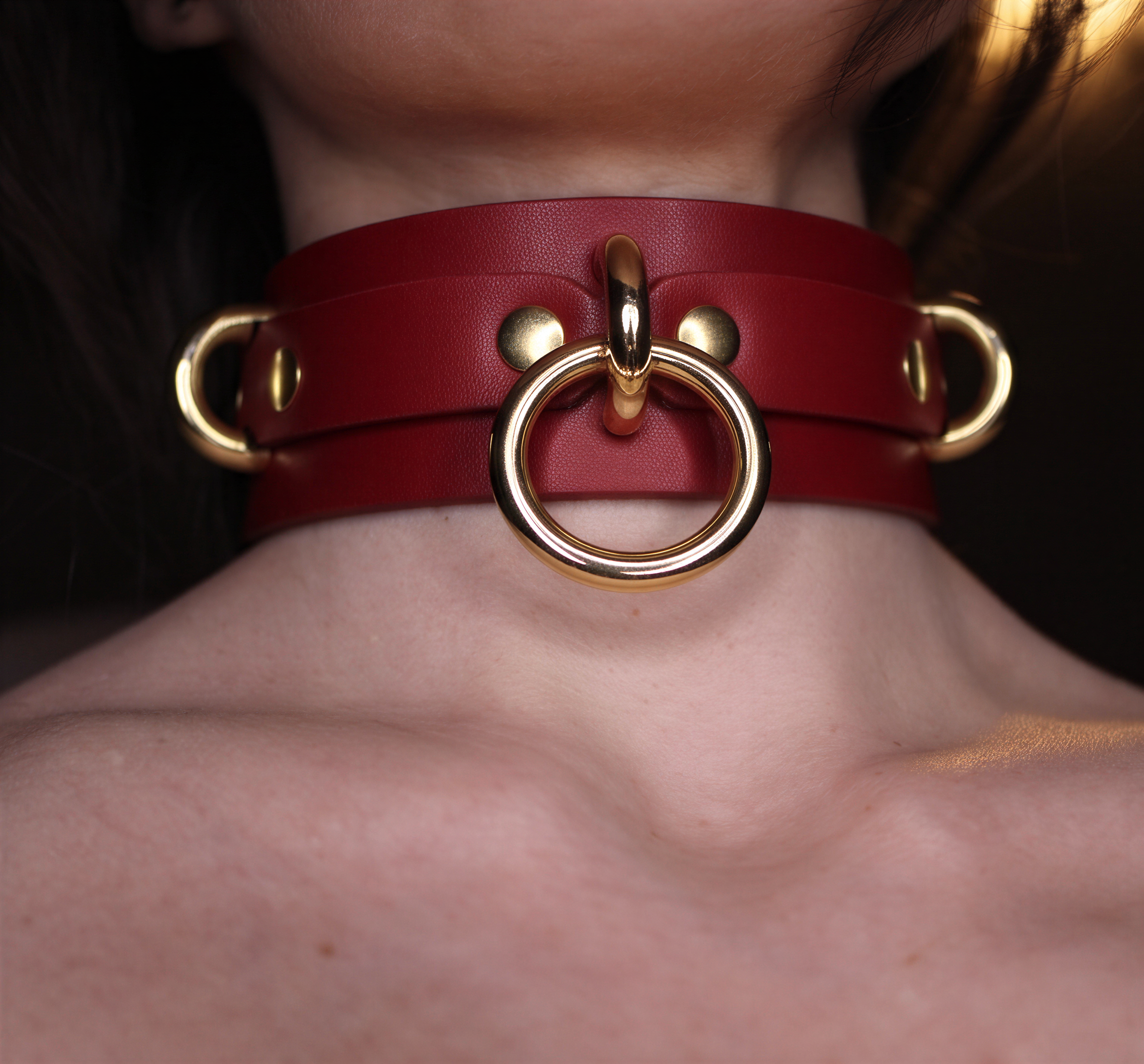 A close-up of a person's neck wearing a red leather choker necklace with gold-colored metal rings.