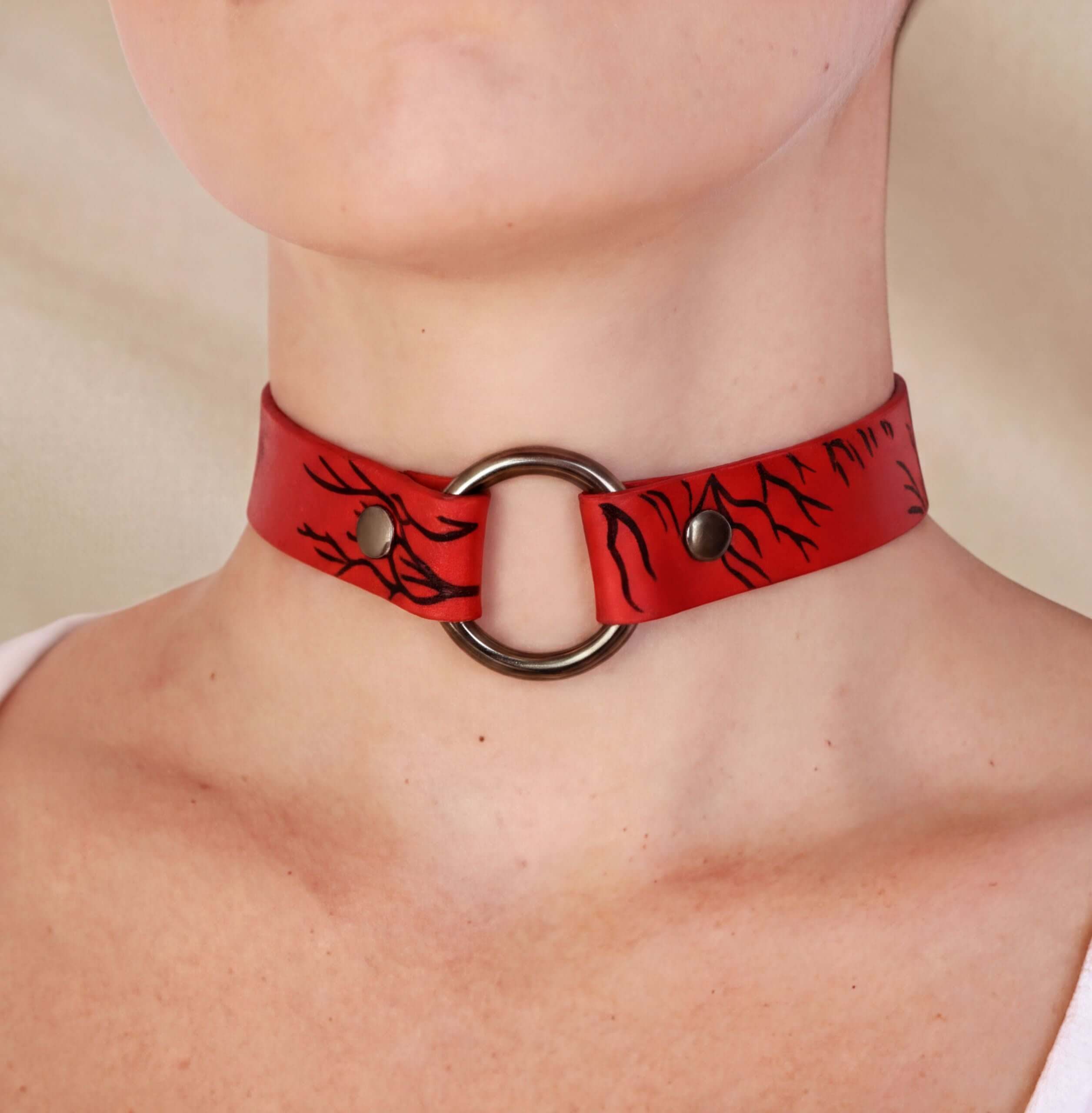 A close-up of a person wearing a red choker necklace with a metal ring and a black leaf design.