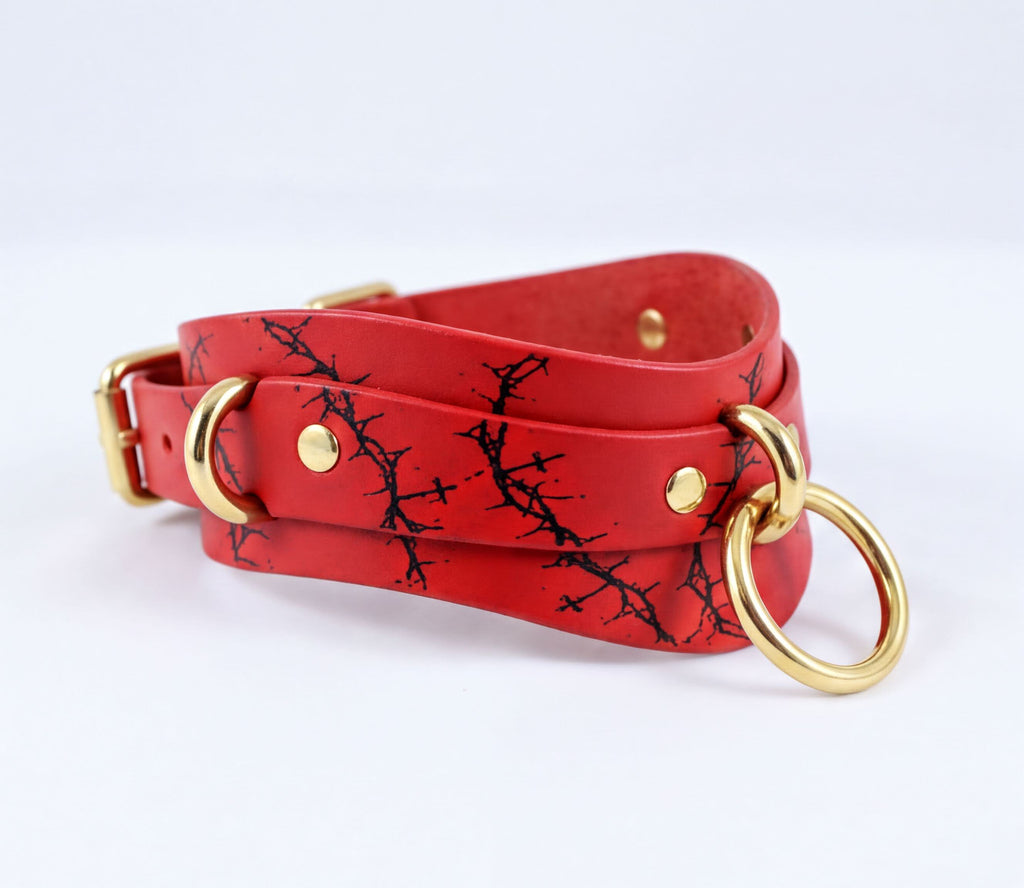 A red leather choker necklace with a black tree branch pattern and gold-colored metal rings.
