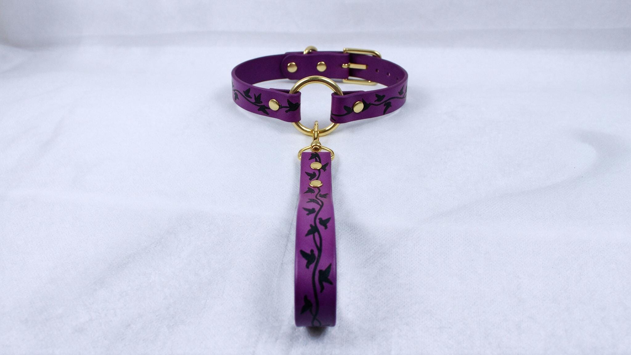 A purple collar with a gold buckle and a black leaf design on the strap is displayed against a white background.