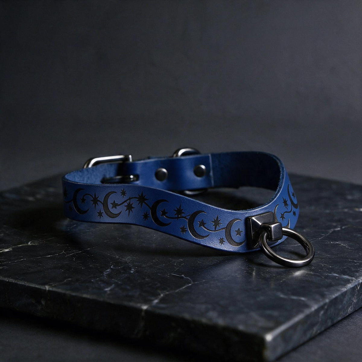 A blue leather collar with a star and crescent design, featuring a metal ring and buckle, is displayed on a dark surface.