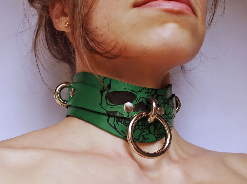 A close-up of a person's neck and chest area wearing a green leather choker necklace with metal rings and a skull design.