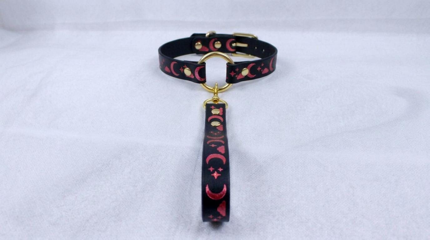 A black collar with a pink and black patterned strap and a gold-colored buckle is displayed on a white surface.