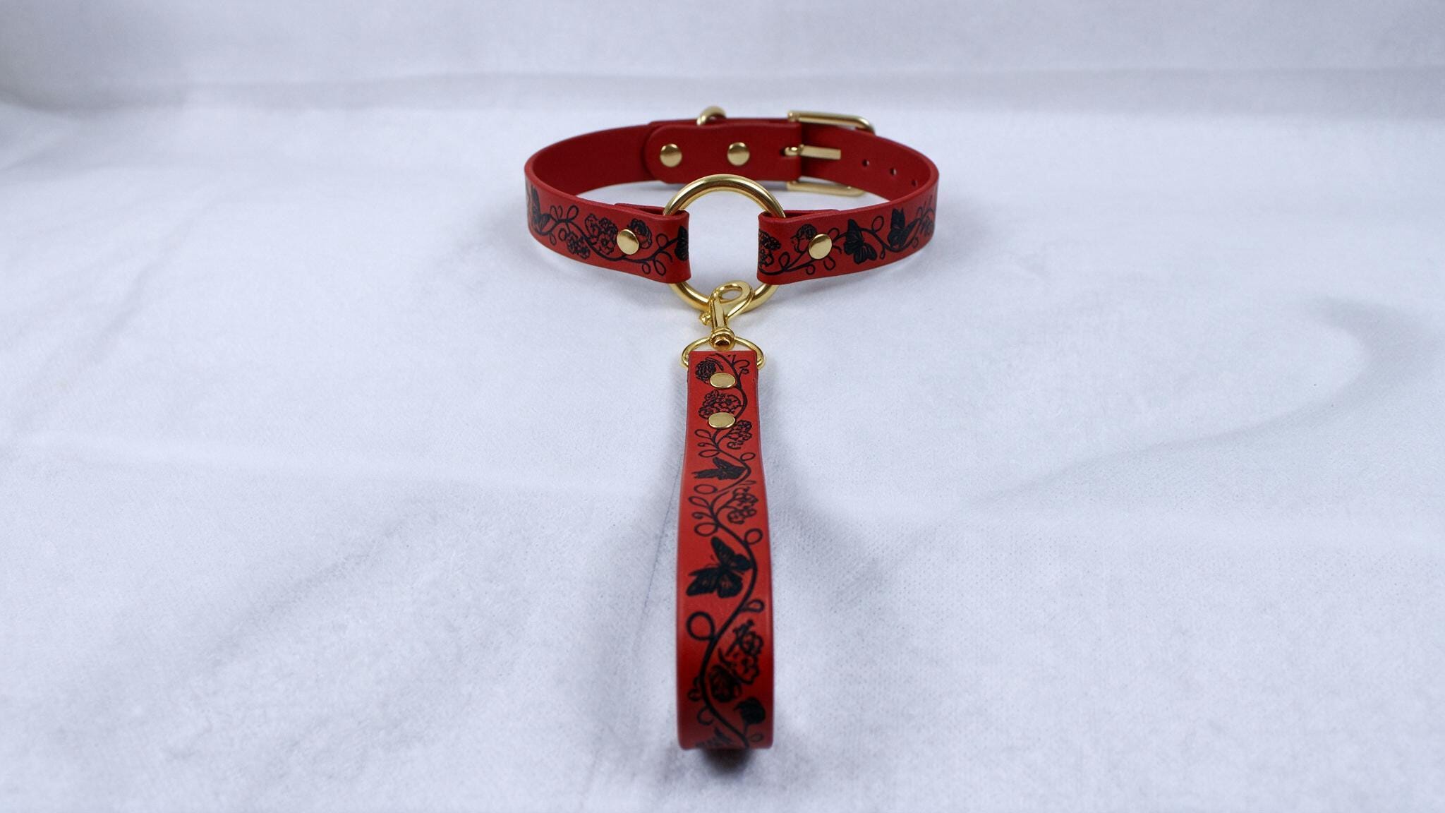 A red leather collar with a gold buckle and a black floral pattern on the strap is displayed on a white surface.