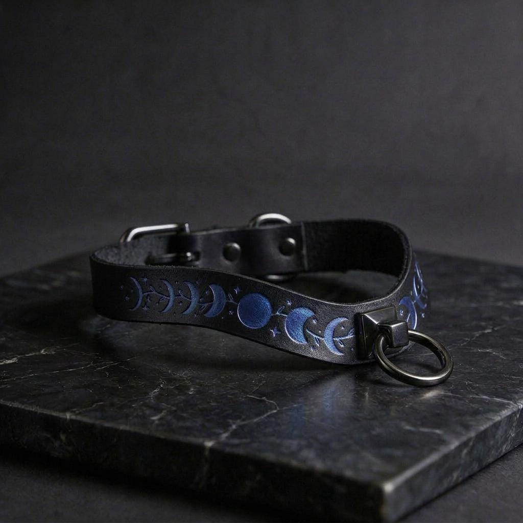 A black leather collar with blue moon and star designs on it, resting on a dark surface.
