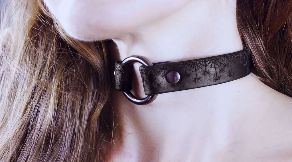 A close-up of a person's neck and chin wearing a black choker necklace with a spider web design.