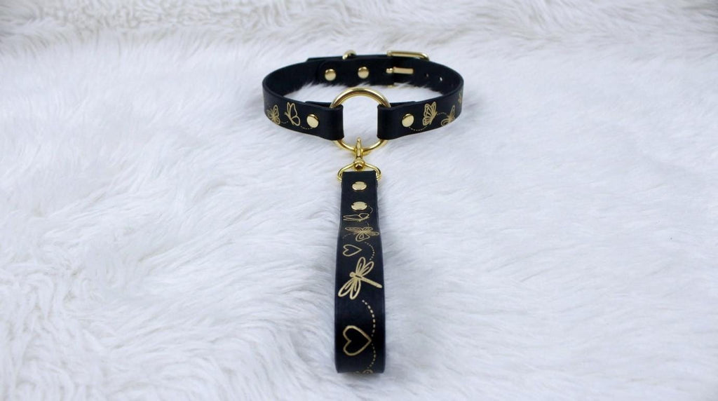 A black leather collar with gold accents and a black handle with gold accents, placed on a white furry surface.