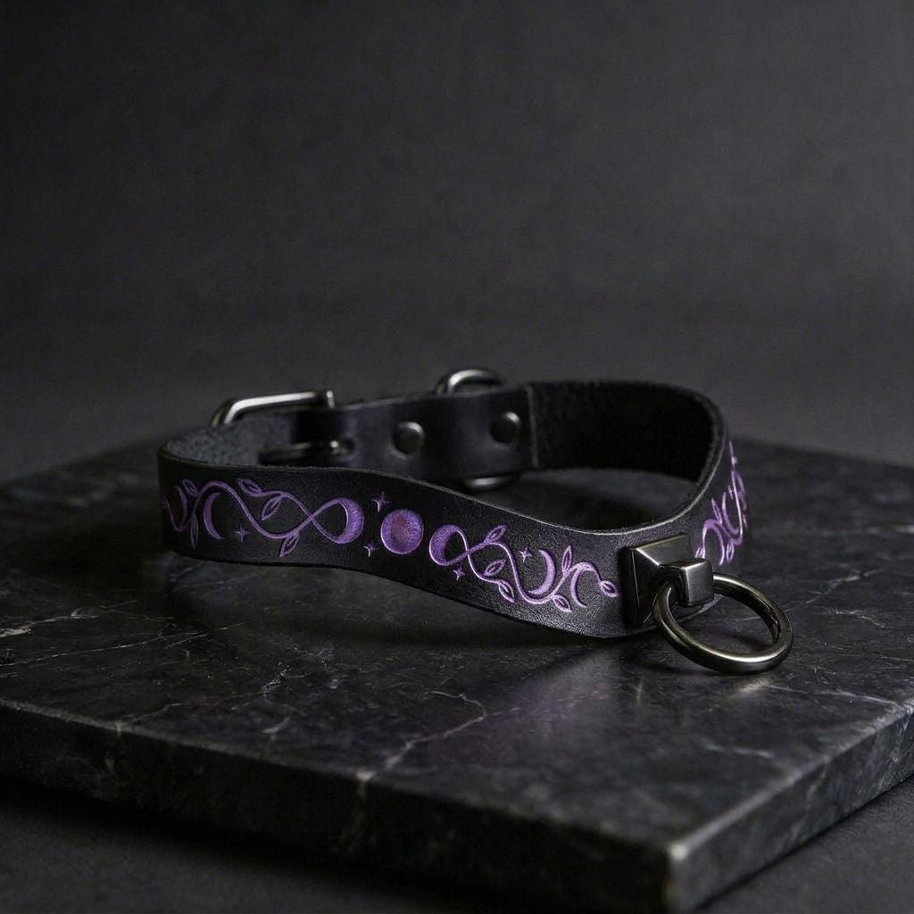 A black leather collar with purple and white designs, including a snake and moon motif, is displayed on a dark surface.