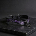 A black leather collar with purple and white designs, including a snake and moon motif, is displayed on a dark surface.